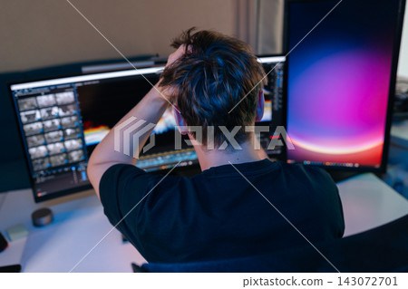 Man feeling stressed and frustrated, gripping head while working on computer with multiple screens in dark home office, experiencing burnout and overwhelming deadlines late at night. 143072701