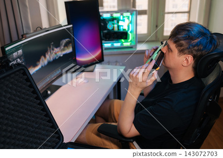 Man in black tee and shorts reclines in modern home office, holding small gadget to nose while inspecting new computer hardware beside rgb lit monitors and desktop pc. Man in black tee and shorts reclines in modern home office, holding small gadget to nose while inspecting new computer hardware beside rgb lit monitors and desktop pc. 143072703
