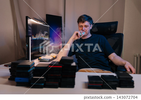 Man in black t shirt vaping electronic cigarette while working on computer surrounded by external hard drives, concept of data storage, technology, contemporary lifestyle. Man in black t shirt vaping electronic cigarette while working on computer surrounded by external hard drives, concept of data storage, technology, contemporary lifestyle. 143072704