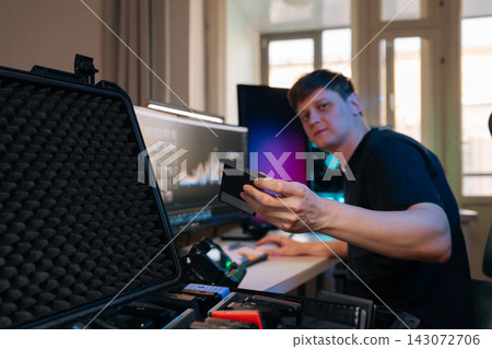 Male creative professional sitting at desk, managing digital files. He holding portable SSD in hand, with open protective case full of storage media in foreground. Modern home office workflow. Male creative professional sitting at desk, managing digital files. He holding portable SSD in hand, with open protective case full of storage media in foreground. Modern home office workflow. 143072706