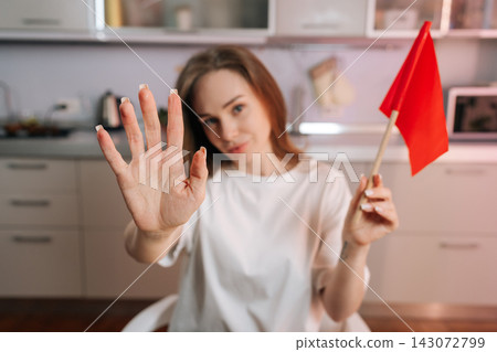 Young woman holds red flag, signaling warning danger. Her hand is in foreground showing stop gesture. Concept for relationship problems, boundaries, dealbreakers. Shallow depth of field at home. Young woman holds red flag, signaling warning danger. Her hand is in foreground showing stop gesture. Concept for relationship problems, boundaries, dealbreakers. Shallow depth of field at home. 143072799