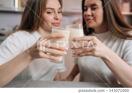 Two young women in white shirts sit at table, toasting with glasses of milk. Shallow depth of field focuses on hands and glassware. Casual, healthy lifestyle concept for nutrition or friendship. Two young women in white shirts sit at table, toasting with glasses of milk. Shallow depth of field focuses on hands and glassware. Casual, healthy lifestyle concept for nutrition or friendship. 143072800