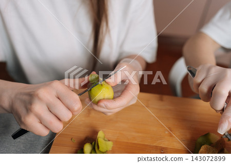 Close-up shot of two people wearing white shirts and disposable gloves preparing fresh kiwi. They are peeling green fruit with knives over wooden cutting board. Concept of healthy eating and hygiene. 143072899