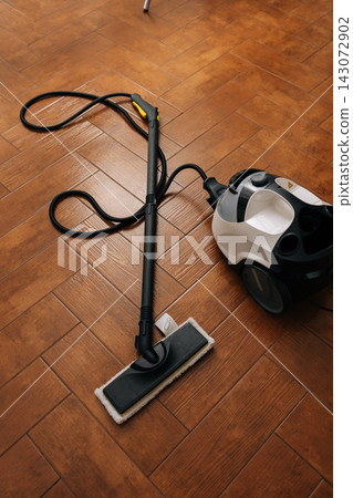 Top view of modern steam cleaner appliance and flat mop attachment resting on warm brown herringbone tile floor, signifying thorough home hygiene, deep cleaning, and efficient floor care. Top view of modern steam cleaner appliance and flat mop attachment resting on warm brown herringbone tile floor, signifying thorough home hygiene, deep cleaning, and efficient floor care. 143072902