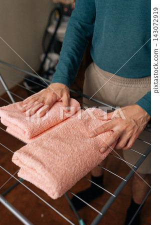 Vertical cropped shot of unrecognizable man folding clean, soft pink towels on laundry drying rack, performing daily household chores and maintaining fresh, organized linens indoors. Vertical cropped shot of unrecognizable man folding clean, soft pink towels on laundry drying rack, performing daily household chores and maintaining fresh, organized linens indoors. 143072919