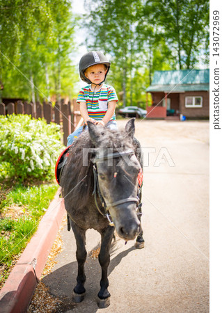 Beautiful little girl two years old riding pony horse in big safety jockey helmet posing outdoors on countryside Beautiful little girl two years old riding pony horse in big safety jockey helmet posing outdoors on countryside 143072969