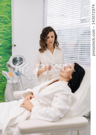 Middle-aged aesthetician doctor applying ultrasound lifting device to female client face during professional facial treatment in modern beauty clinic for skin rejuvenation and anti-aging care. Middle-aged aesthetician doctor applying ultrasound lifting device to female client face during professional facial treatment in modern beauty clinic for skin rejuvenation and anti-aging care. 143072974