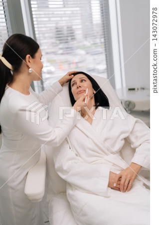 Vertical portrait of middle-aged female client consulting with medical aesthetic specialist who marking facial contours and treatment areas with white pencil during pre-procedure planning in clinic. Vertical portrait of middle-aged female client consulting with medical aesthetic specialist who marking facial contours and treatment areas with white pencil during pre-procedure planning in clinic. 143072978