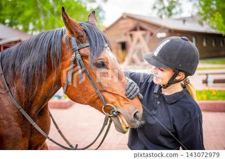 Lovely young woman wearing helmet stroking to her brown horse Lovely young woman wearing helmet stroking to her brown horse 143072979