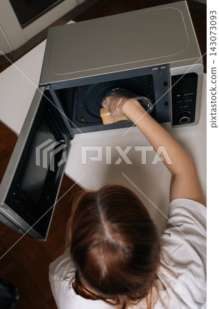 High-angle view of housewife in clear disposable gloves scrubbing inside of microwave with yellow sponge, close-up of routine kitchen cleaning and appliance hygiene maintenance. High-angle view of housewife in clear disposable gloves scrubbing inside of microwave with yellow sponge, close-up of routine kitchen cleaning and appliance hygiene maintenance. 143073093