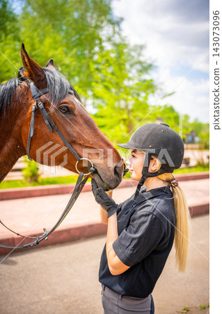 Lovely young woman wearing helmet stroking to her brown horse Lovely young woman wearing helmet stroking to her brown horse 143073096