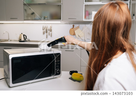 Back view of young woman using handheld steam cleaner with white scrubbing pad for deep cleaning top of microwave oven in modern kitchen, ensuring proper hygiene. Concept of household chores. Back view of young woman using handheld steam cleaner with white scrubbing pad for deep cleaning top of microwave oven in modern kitchen, ensuring proper hygiene. Concept of household chores. 143073103