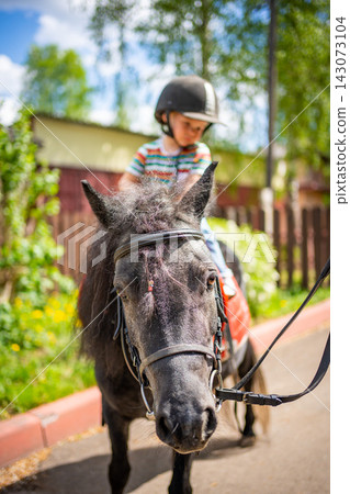 Beautiful little girl two years old riding pony horse in big safety jockey helmet posing outdoors on countryside  143073104