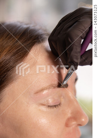 Close up of eyebrow hair removal with tweezers by gloved hand on female face profile Close up of eyebrow hair removal with tweezers by gloved hand on female face profile 143073125