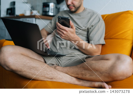 Casual man sits cross legged on bright orange sofa, multitasking with laptop and smartphone. Concept of remote work, digital nomad life, connectivity, and modern lifestyle. Close-up shot. Casual man sits cross legged on bright orange sofa, multitasking with laptop and smartphone. Concept of remote work, digital nomad life, connectivity, and modern lifestyle. Close-up shot. 143073278