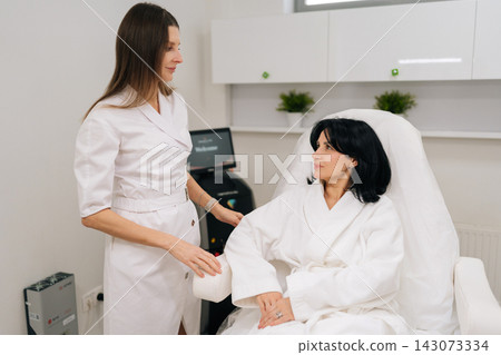 Cosmetologist in white uniform consulting middle-aged client in robe, discussing personalized facial and antiaging treatment plan in modern dermatology spa with advanced equipment. Cosmetologist in white uniform consulting middle-aged client in robe, discussing personalized facial and antiaging treatment plan in modern dermatology spa with advanced equipment. 143073334