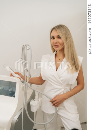 Vertical portrait of blonde esthetician in white coat setting up modern fractional radiofrequency microneedling device, adjusting parameters before facial rejuvenation therapy, looking at camera. 143073348