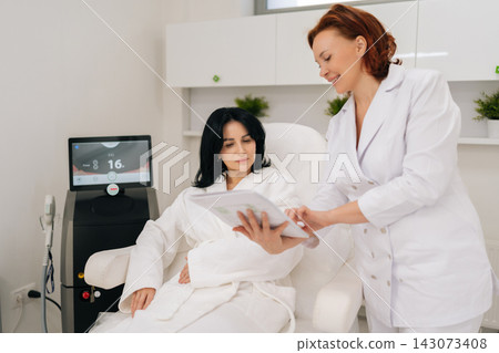 Positive cosmetologist in uniform talking with female client in bathrobe seated in treatment chair, reviewing information on document before cosmetic procedure using advanced technology apparatus. 143073408