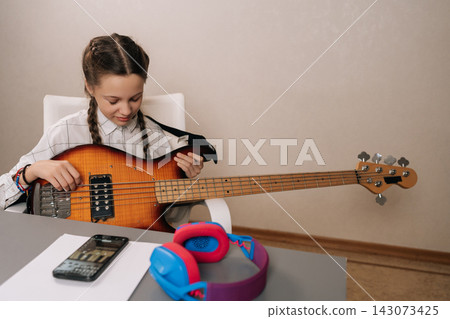Portrait of cute preteen girl with pigtails focused on playing bass guitar, following online lesson on smartphone, developing new musical skills and a creative hobby at home. Portrait of cute preteen girl with pigtails focused on playing bass guitar, following online lesson on smartphone, developing new musical skills and a creative hobby at home. 143073425