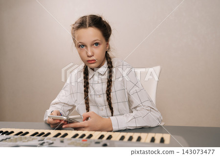 Cute preteen girl pianist with pigtails sitting at electronic piano, focused on smartphone instead of music practice, illustrating modern screen time distraction during home lesson, looking at camera. Cute preteen girl pianist with pigtails sitting at electronic piano, focused on smartphone instead of music practice, illustrating modern screen time distraction during home lesson, looking at camera. 143073477