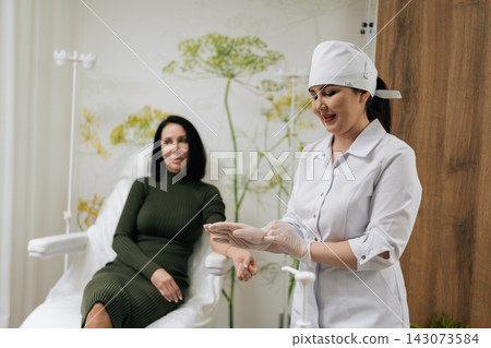 Female medical worker pulling on sterile examination glove, ensuring safety and hygiene before medical procedure with blurred patient receiving care in background. Concept of healthy lifestyle. Female medical worker pulling on sterile examination glove, ensuring safety and hygiene before medical procedure with blurred patient receiving care in background. Concept of healthy lifestyle. 143073584