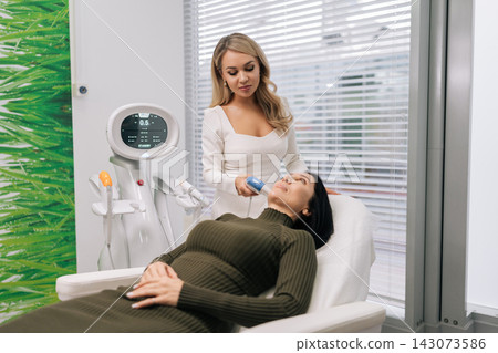 Brunette middle-aged woman receiving advanced aesthetic procedure using ultrasound device for facial skin lifting and rejuvenation, improving elasticity and youthfulness in beauty clinic. Brunette middle-aged woman receiving advanced aesthetic procedure using ultrasound device for facial skin lifting and rejuvenation, improving elasticity and youthfulness in beauty clinic. 143073586