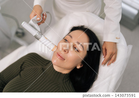 Close-up portrait of female getting non-surgical aesthetic procedure, cosmetologist applying ultrasound device to face, stimulating collagen production for skin tightening and youthful appearance. Close-up portrait of female getting non-surgical aesthetic procedure, cosmetologist applying ultrasound device to face, stimulating collagen production for skin tightening and youthful appearance. 143073591