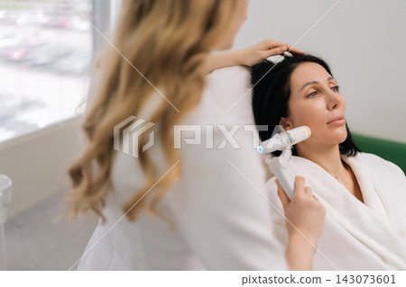 Rear view of female cosmetologist performing rejuvenating ultrasound lifting facial treatment on adult woman face at modern aesthetic clinic, focusing on anti-aging and skin contouring. Rear view of female cosmetologist performing rejuvenating ultrasound lifting facial treatment on adult woman face at modern aesthetic clinic, focusing on anti-aging and skin contouring. 143073601