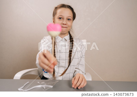 Studio shot of cheerful young girl smiling looking at camera, holding giving pastel pink and white heart shaped lollipop, representing innocence and childhood joy. Studio shot of cheerful young girl smiling looking at camera, holding giving pastel pink and white heart shaped lollipop, representing innocence and childhood joy. 143073673
