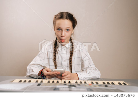 Portrait of joyful young girl with pigtails showing playful expression and sticking out tongue, enjoying music lessons with a modern electronic keyboard and digital device. Portrait of joyful young girl with pigtails showing playful expression and sticking out tongue, enjoying music lessons with a modern electronic keyboard and digital device. 143073676
