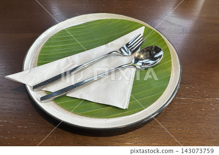 top view cutlery placed on a triangular folded paper napkin, set on a banana leaf atop a white ceramic plate, resting on a wooden floor background, object, decor, copy space 143073759