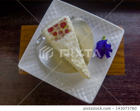 top view sweet triangular slice of white cake resting on a circular metallic silver paper tray and butterfly pea flower on a white ceramic plate on wooden background, food, dessert, copy space 143073760
