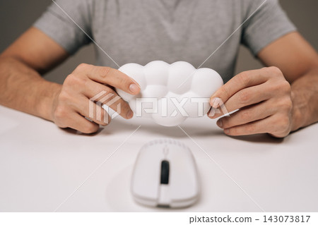 Close-up of man holding cloud-shaped wrist rest for computer mouse, providing ergonomic support and comfort working on home office desk, preventing carpal tunnel syndrome and repetitive strain injury. Close-up of man holding cloud-shaped wrist rest for computer mouse, providing ergonomic support and comfort working on home office desk, preventing carpal tunnel syndrome and repetitive strain injury. 143073817
