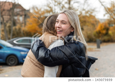 Smiling woman hugs friend outdoors during candid autumn meeting, expressing joy, affection and emotional connection. Urban background and fall colors suggest reunion, greeting or emotional goodbye Smiling woman hugs friend outdoors during candid autumn meeting, expressing joy, affection and emotional connection. Urban background and fall colors suggest reunion, greeting or emotional goodbye 143073939