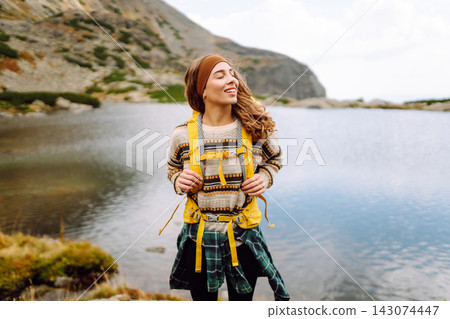 Hiking near a lake in mountain area with a woman wearing a yellow jacket and backpack during daytime Hiking near a lake in mountain area with a woman wearing a yellow jacket and backpack during daytime 143074447