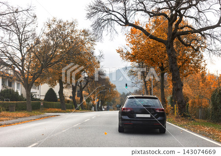Back scenic view black family car driving wet countryside road through bright autumnal golden red orange nature foliage forest trees. Scenery fall season highway vehicle. Drive auto safety Back scenic view black family car driving wet countryside road through bright autumnal golden red orange nature foliage forest trees. Scenery fall season highway vehicle. Drive auto safety 143074669