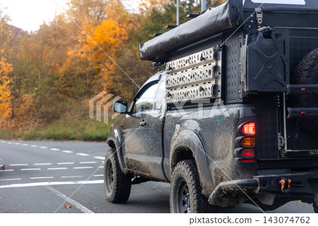 Rugged pickup with expedition camper waits on wet autumn road beside glowing forest, suggesting outdoor escape. Muddy vehicle, travel gear and fall colors evoke adventure, travel and freedom Rugged pickup with expedition camper waits on wet autumn road beside glowing forest, suggesting outdoor escape. Muddy vehicle, travel gear and fall colors evoke adventure, travel and freedom 143074762