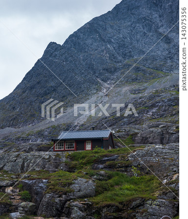 Red wooden cabin beneath steep mountain slope, Norway alpine valley Red wooden cabin beneath steep mountain slope, Norway alpine valley 143075356