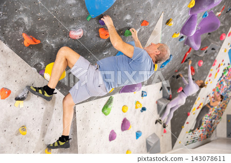 Concentrated elderly man moving on bouldering wall in climbing gym Concentrated elderly man moving on bouldering wall in climbing gym 143078611
