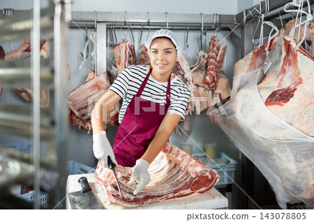 Positive female butcher cutting veal ribs in cold storage room of store Positive female butcher cutting veal ribs in cold storage room of store 143078805