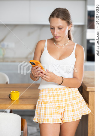 Positive young girl looking at smartphone while standing in kitchen Positive young girl looking at smartphone while standing in kitchen 143079036