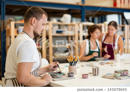 Male potter learns how to make designs on clay plate using brush and paint in pottery workshop Male potter learns how to make designs on clay plate using brush and paint in pottery workshop 143079045