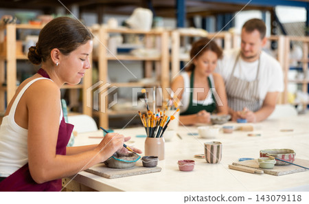 Girl in apron designing handmade clay plate with brush in taller room Girl in apron designing handmade clay plate with brush in taller room 143079118