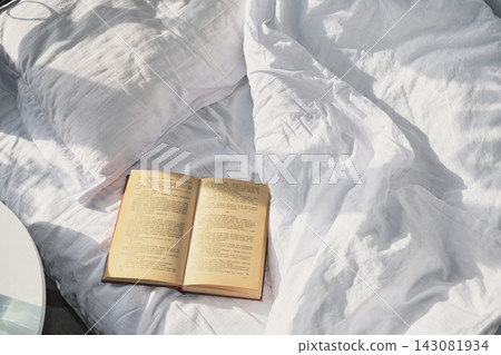 Open book resting on rumpled white linen sheets and pillow in morning light Open book resting on rumpled white linen sheets and pillow in morning light 143081934