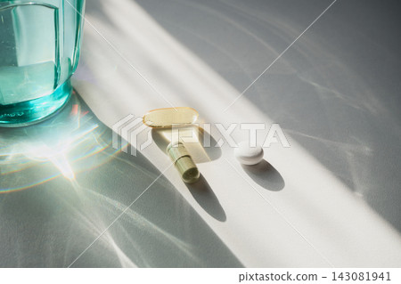 Pills and capsules resting on white surface with glass of water and sunlight Pills and capsules resting on white surface with glass of water and sunlight 143081941