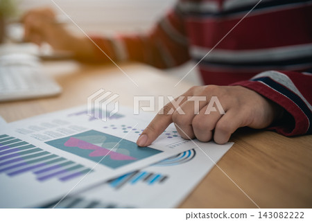 Person analyzing business chart report data with finger pointing on graph paper on wooden desk in office workspace 143082222