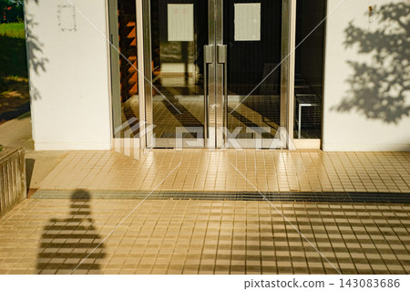The entrance to this modern building features double glass doors and a tiled floor, with the shadows of trees stretching across it. The entrance to this modern building features double glass doors and a tiled floor, with the shadows of trees stretching across it. 143083686