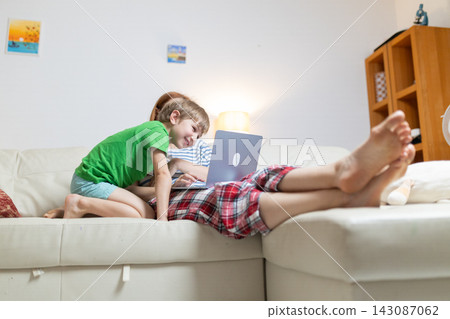 Two children enjoy computer time together while sitting on the couch in a cozy living room during the day Two children enjoy computer time together while sitting on the couch in a cozy living room during the day 143087062