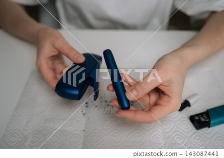 Top view of hands disassembling blue e-cigarette over paper towel with dark residue, showing detailed cleaning and maintenance of heat-not-burn vaping device for hygiene and upkeep. Top view of hands disassembling blue e-cigarette over paper towel with dark residue, showing detailed cleaning and maintenance of heat-not-burn vaping device for hygiene and upkeep. 143087441