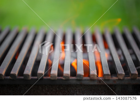 Close-up view of a metal grill with flames visible beneath the slats, set against a blurred green background, highlighting outdoor cooking equipment Close-up view of a metal grill with flames visible beneath the slats, set against a blurred green background, highlighting outdoor cooking equipment 143087576